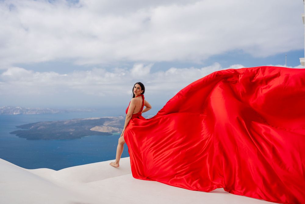 Billowing red dress catches wind on white rooftop overlooking Mediterranean waters.