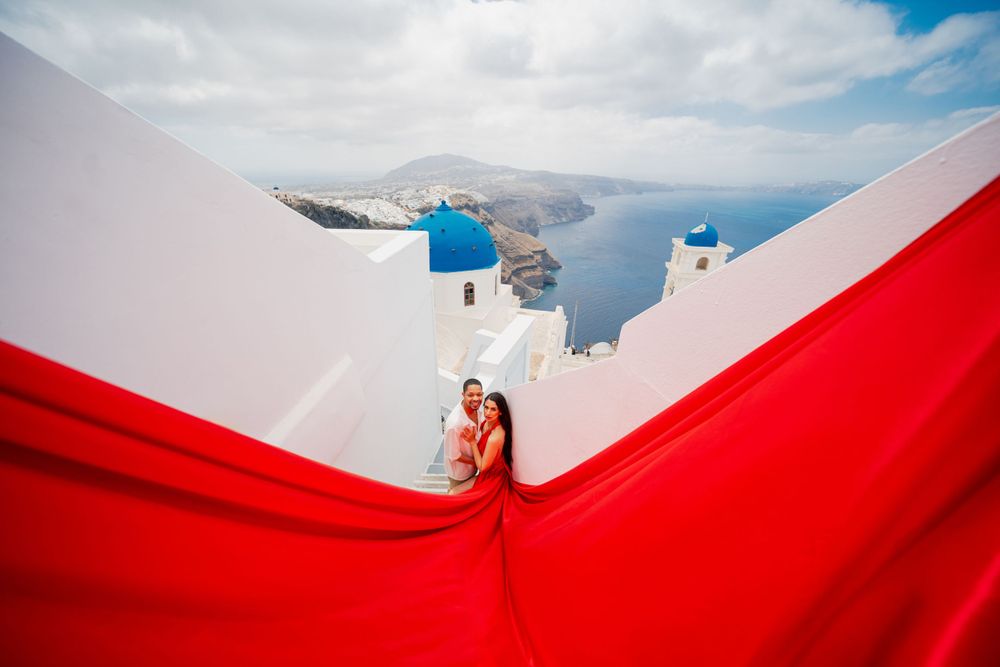 Red dress flows dramatically against white buildings and blue domed church in Santorini.