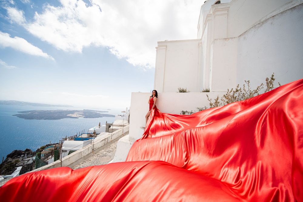 Dramatic red dress fabric billows against classic white Santorini architecture and sea views.
