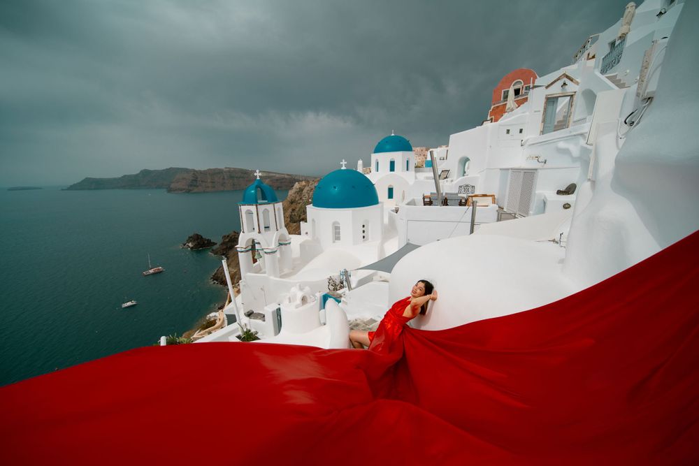 Red flowing fabric dramatically sweeps across white architecture and blue domes in Santorini.