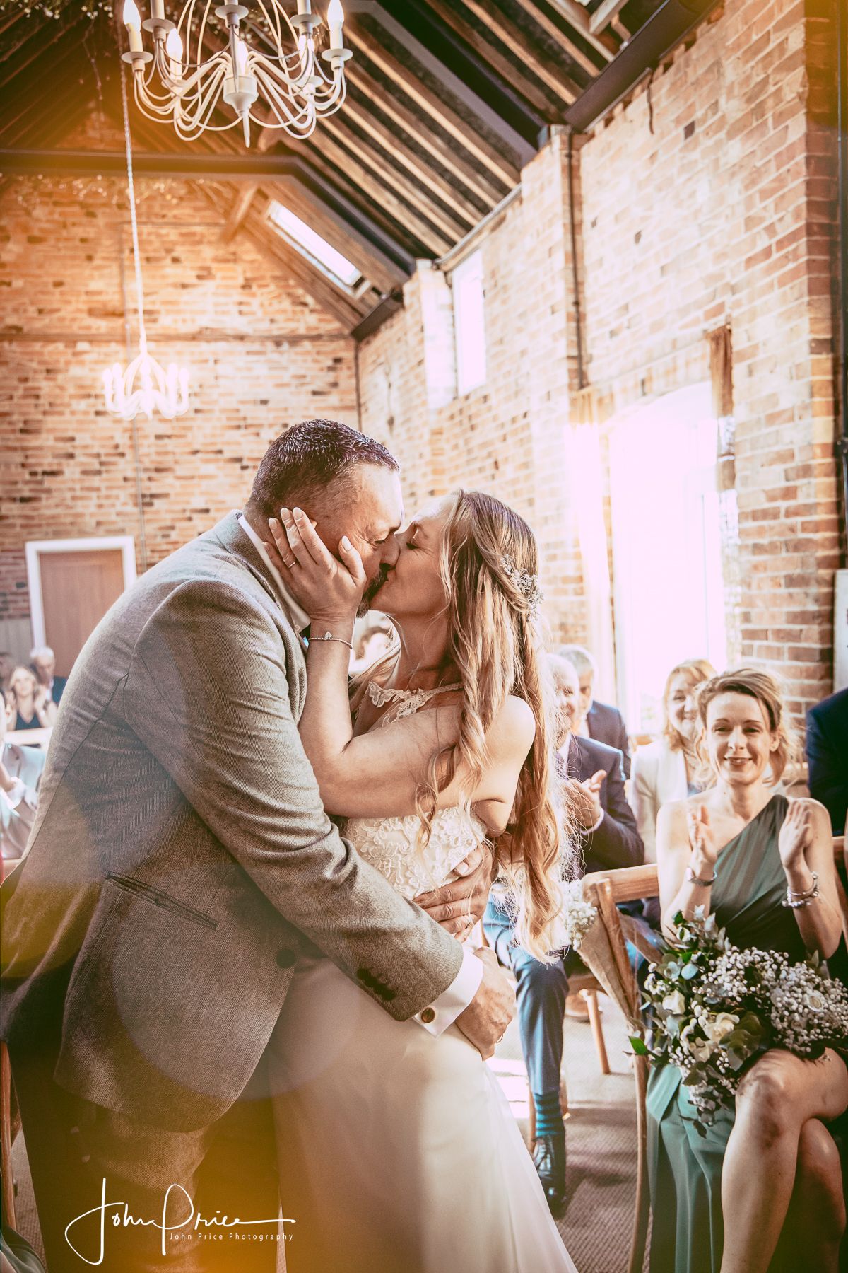 Romantic kiss in a sun-lit rustic brick venue with chandelier overhead.