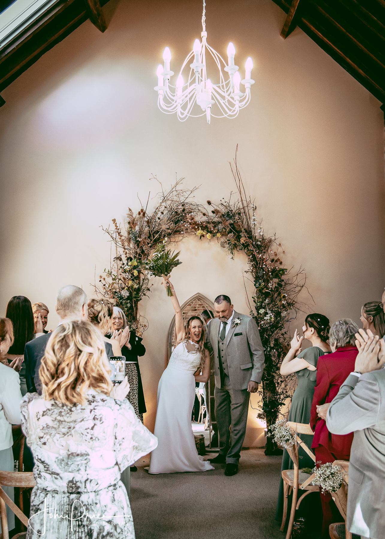 A wedding ceremony in a chapel under an elegant chandelier with a floral arch and guests seated on both sides.