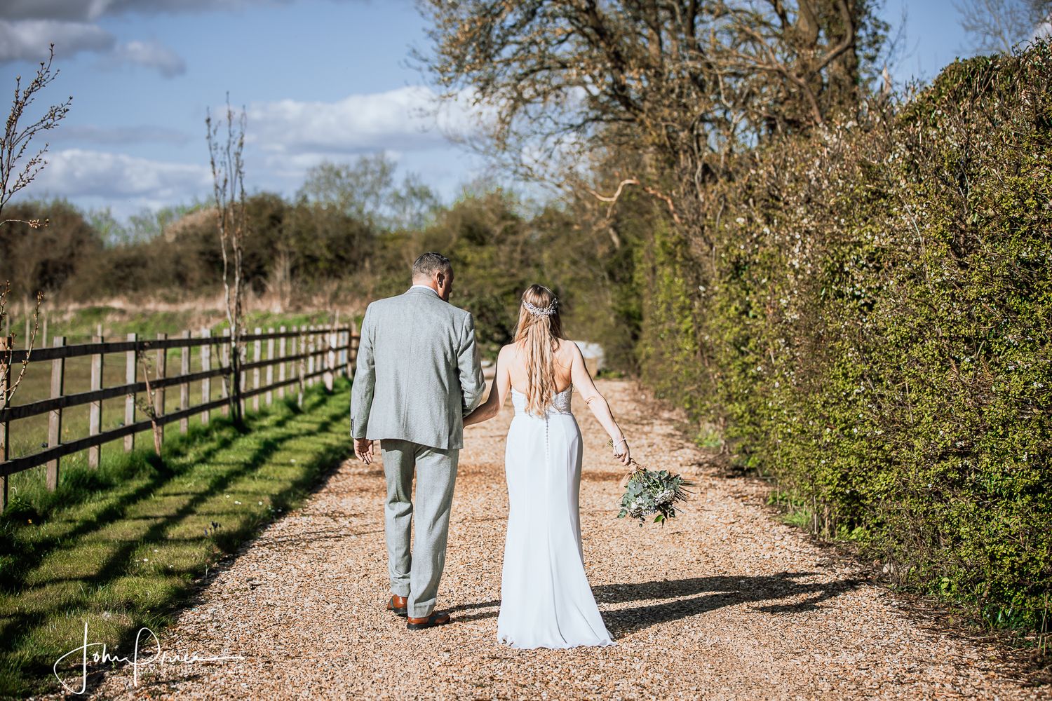 Couple walking hand in hand down a gravel path bordered by wooden fencing.