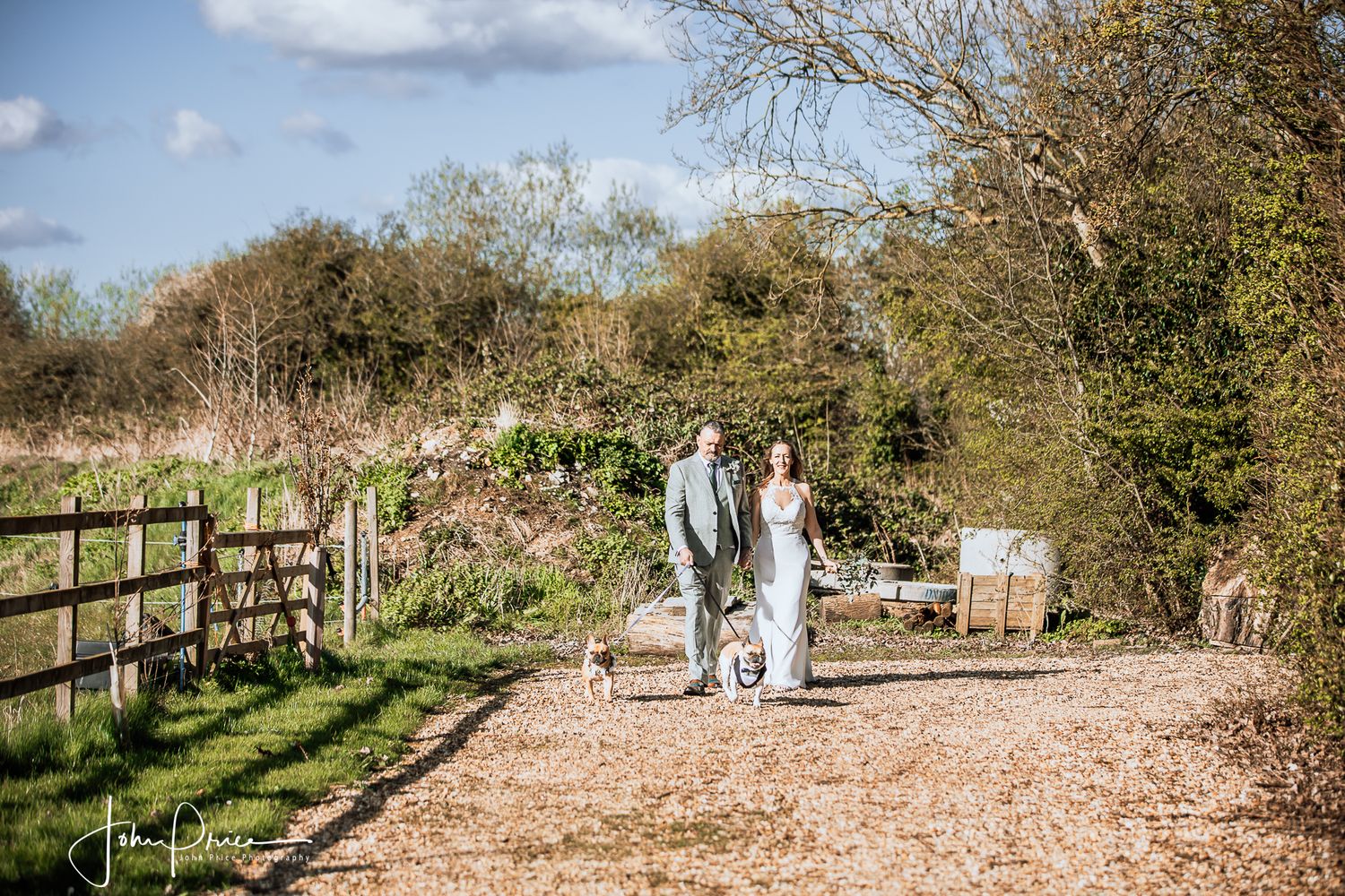 Newlyweds walking down a rustic gravel path bordered by wooden fencing.