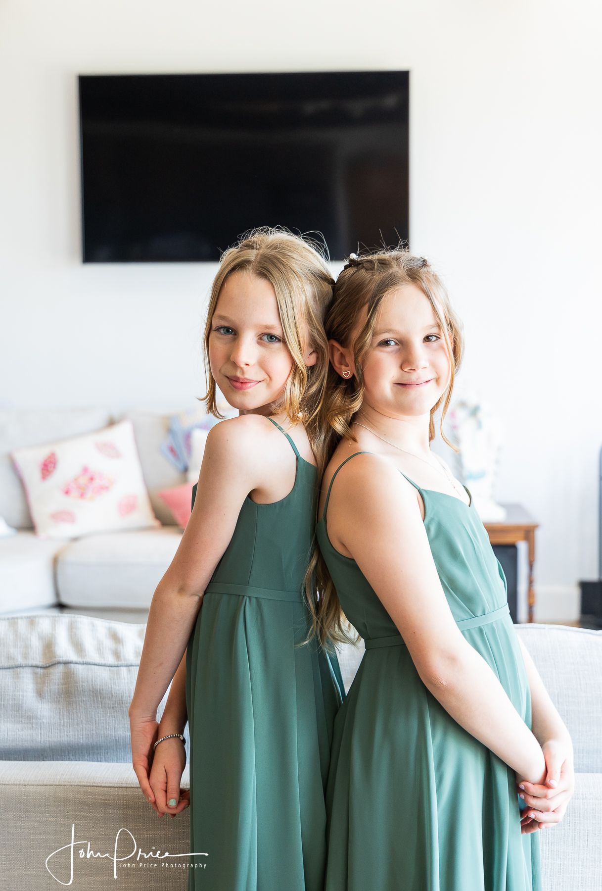 Two young bridesmaids in matching sage green dresses pose back-to-back.