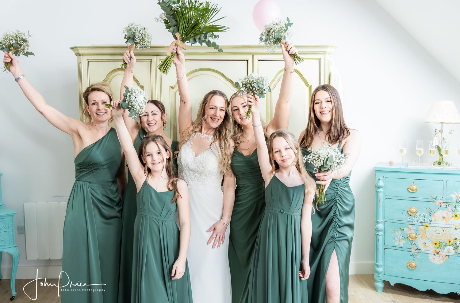 Wedding party poses joyfully in sage green dresses in a bright white room.