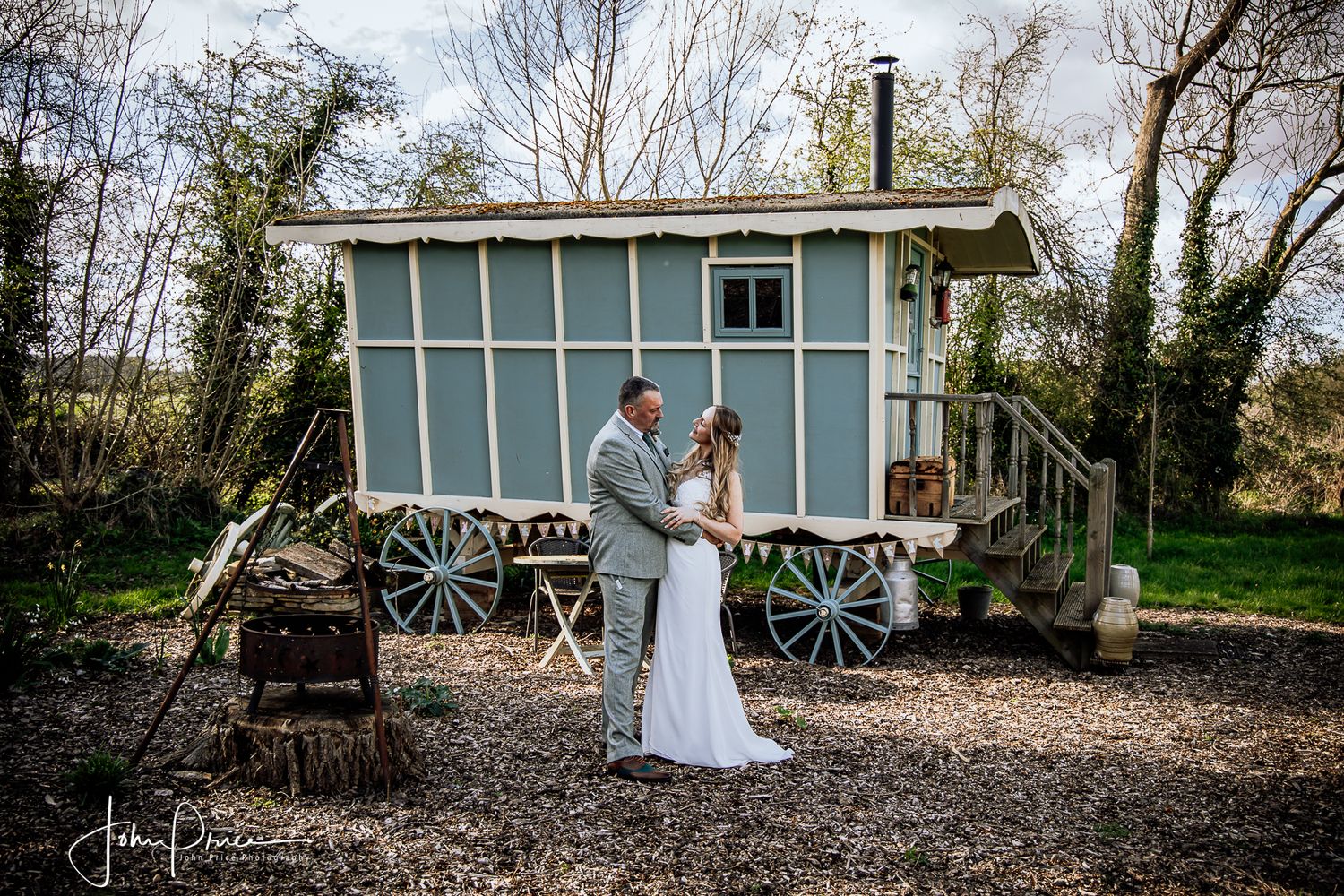 Couple standing in front of a vintage blue shepherd's hut in a rustic outdoor setting.