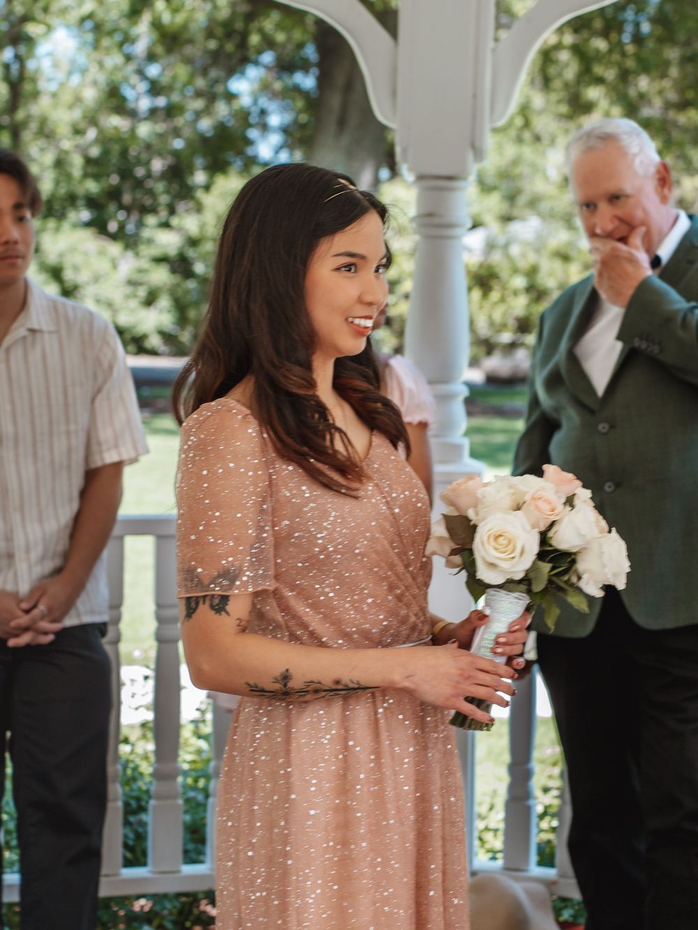 Guest in sparkly rose gold dress holding white rose bouquet at garden wedding ceremony.