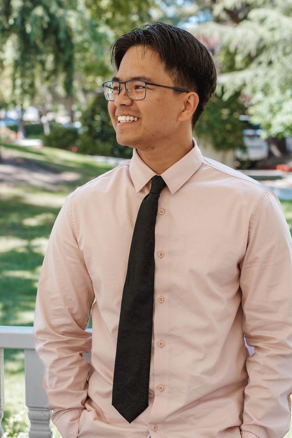 Professional portrait of someone in business attire wearing glasses and a black tie smiling outdoors.