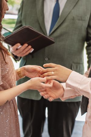 Close up of hands exchanging rings during wedding ceremony with officiant holding book in background.