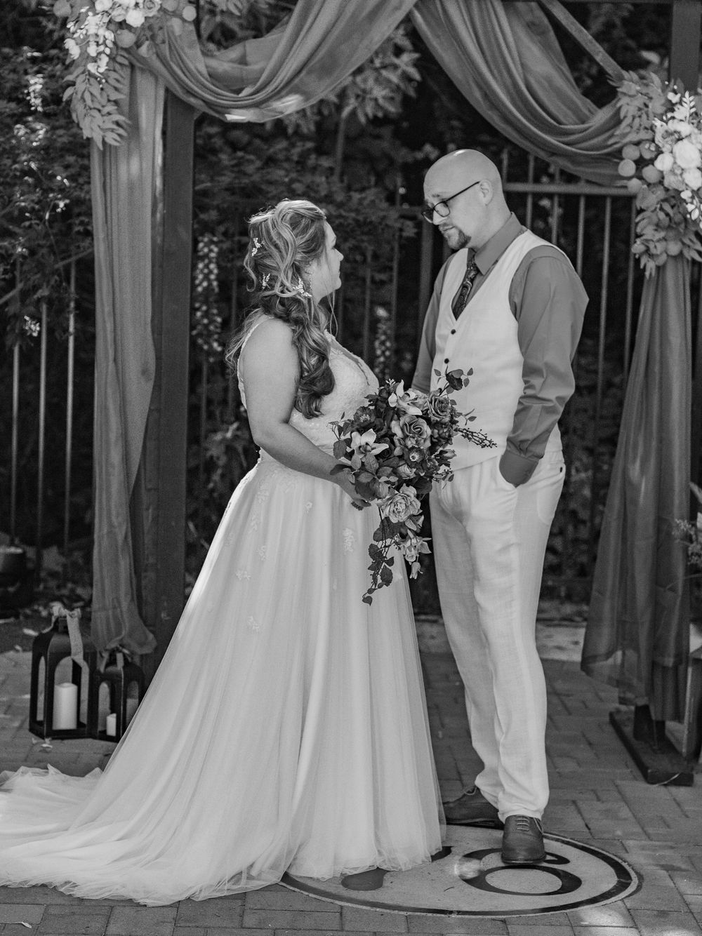 Black and white wedding photo of couple under draped arch with floral decorations.
