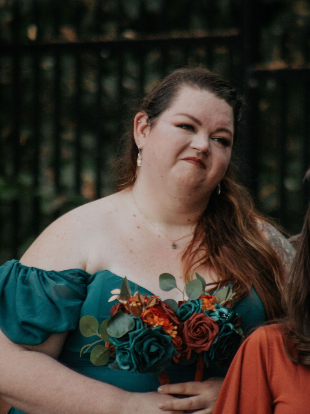 Wedding guest in emerald green dress holding autumn-colored floral bouquet.