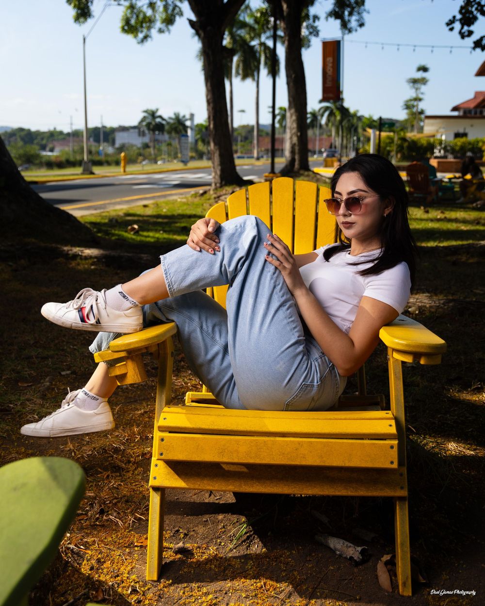 A person relaxes on a bright yellow Adirondack chair in a sunny outdoor setting with palm trees in the background.