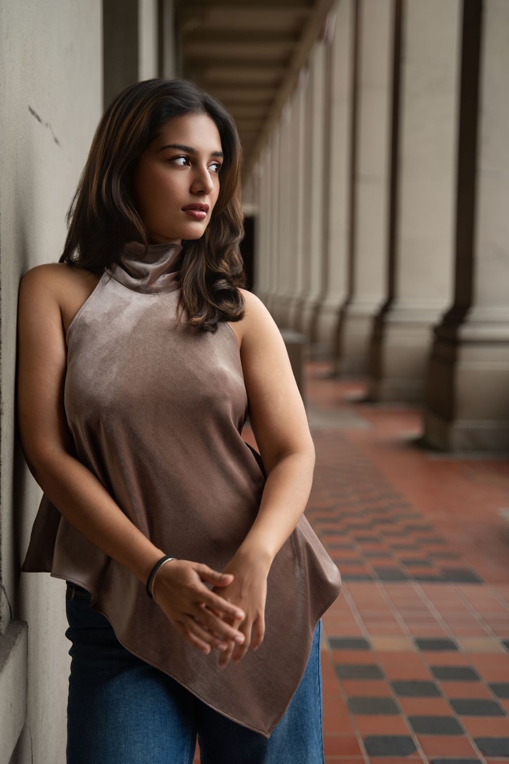 Woman in brown sleeveless top and jeans poses thoughtfully in a historic corridor with checkered floor tiles.