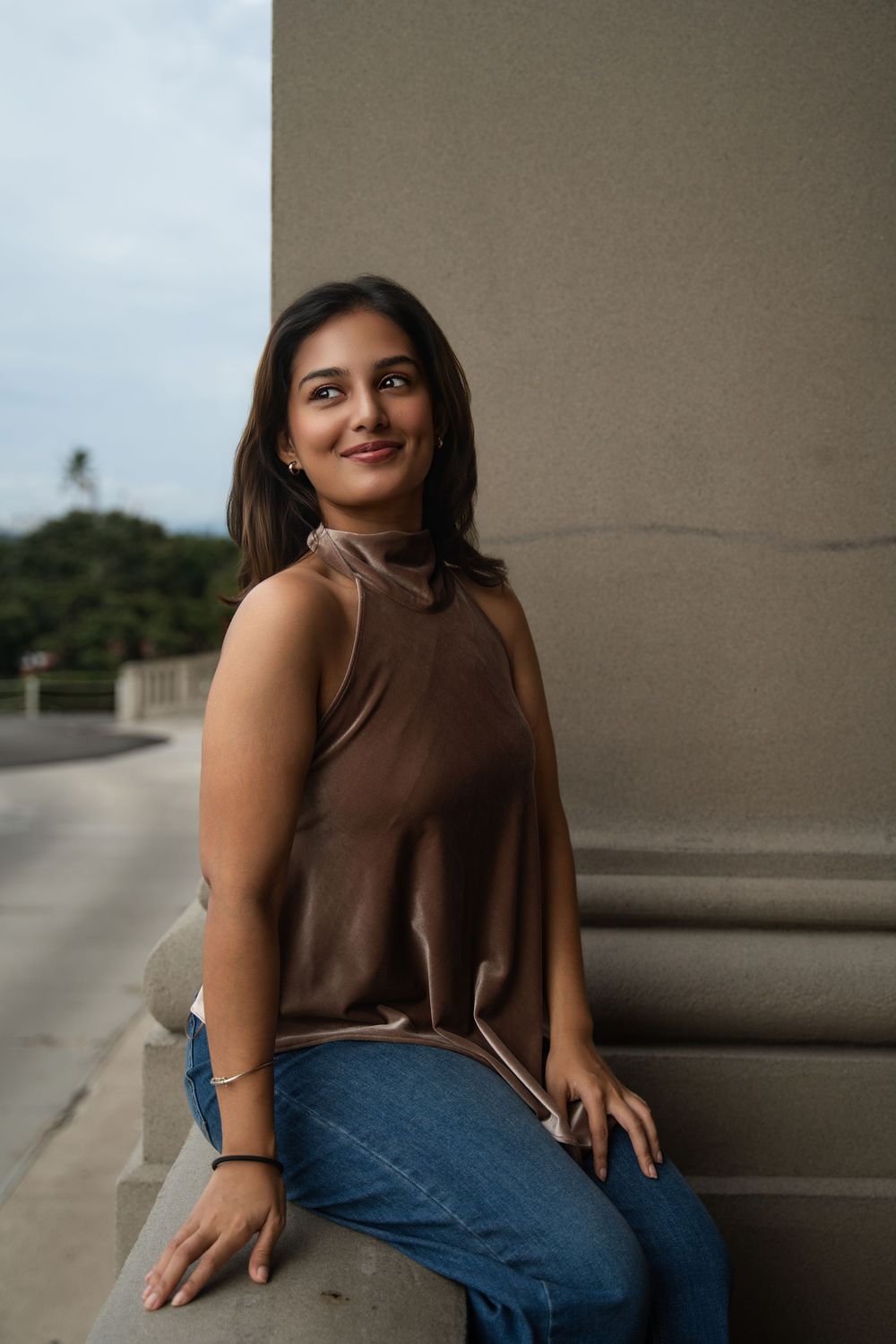 A person in a brown sleeveless top and blue jeans poses casually against a concrete pillar outdoors.