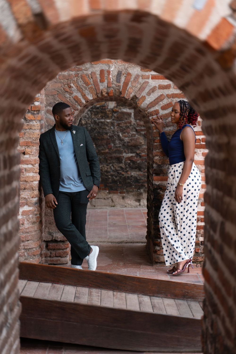 Fashionable couple poses under brick archway in historic building with rustic architectural details.