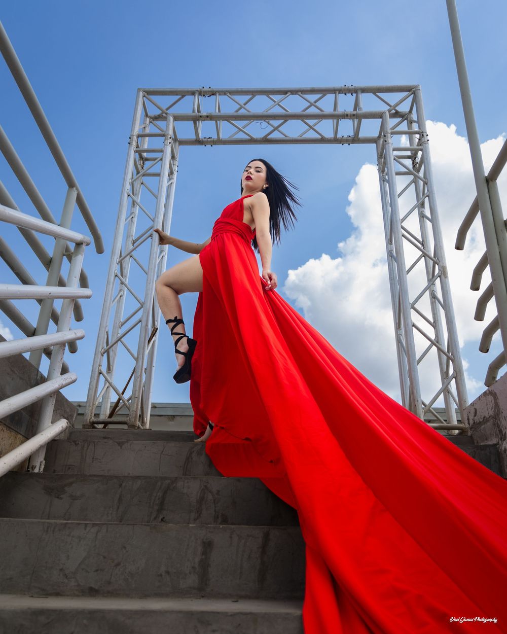 A vibrant red dress billows dramatically against concrete and metal architectural elements under blue sky.