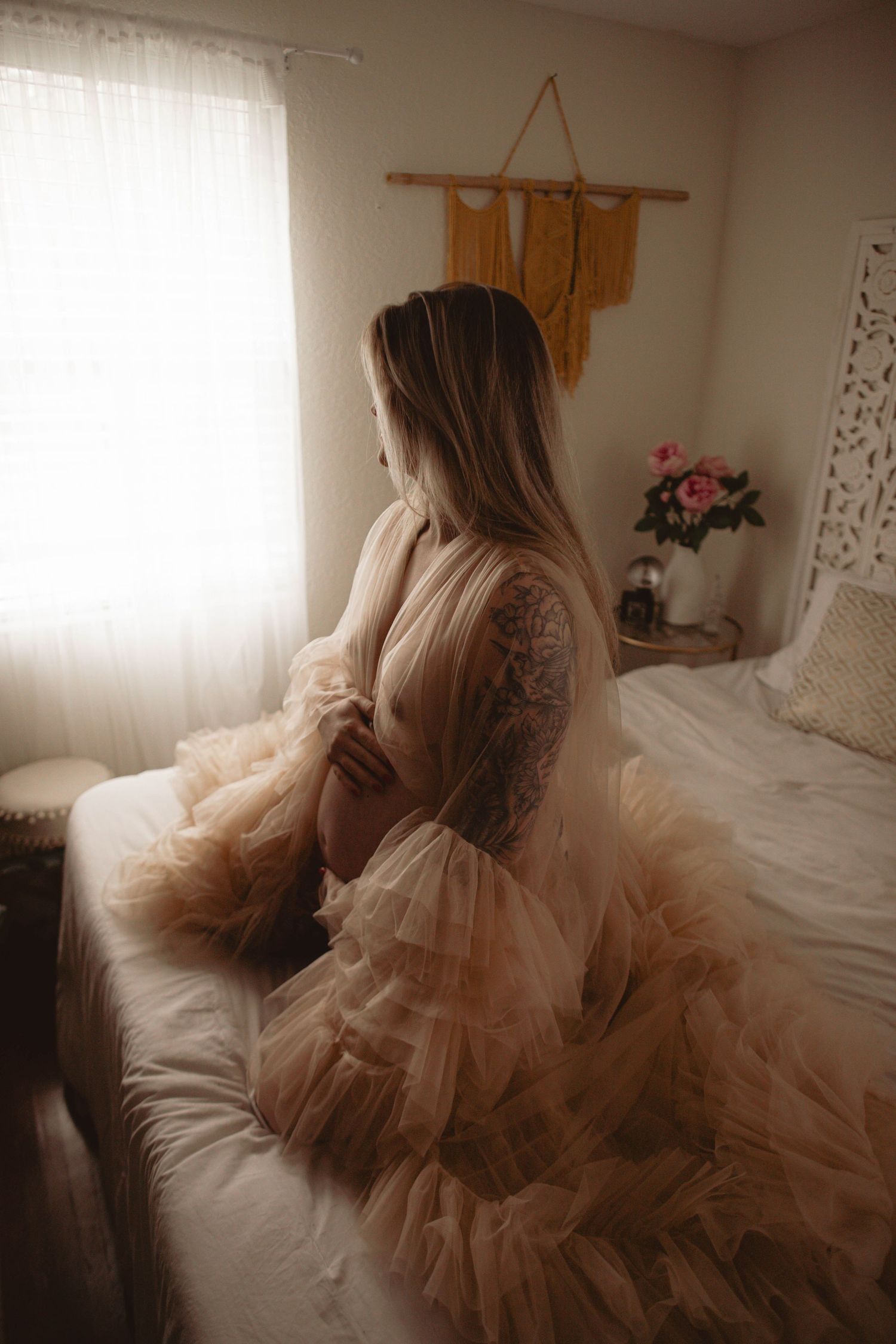 A dreamy boudoir scene with a ruffled peach tulle gown on a white bed near a macrame wall hanging and flowers.
