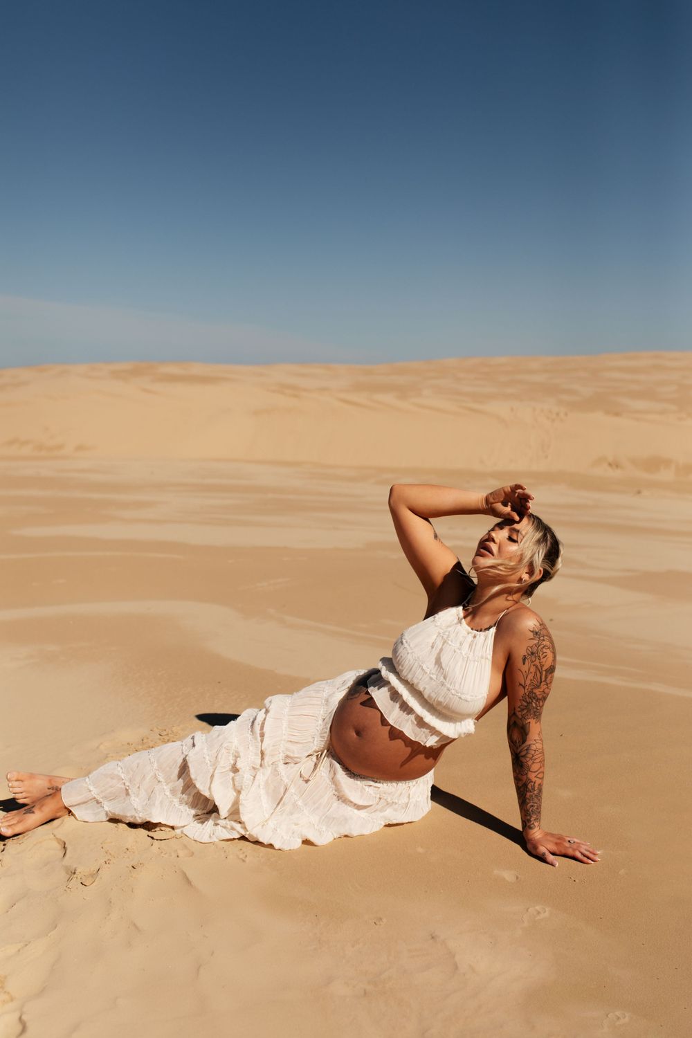 Woman in white dress posing dramatically on desert sand dunes under blue sky.