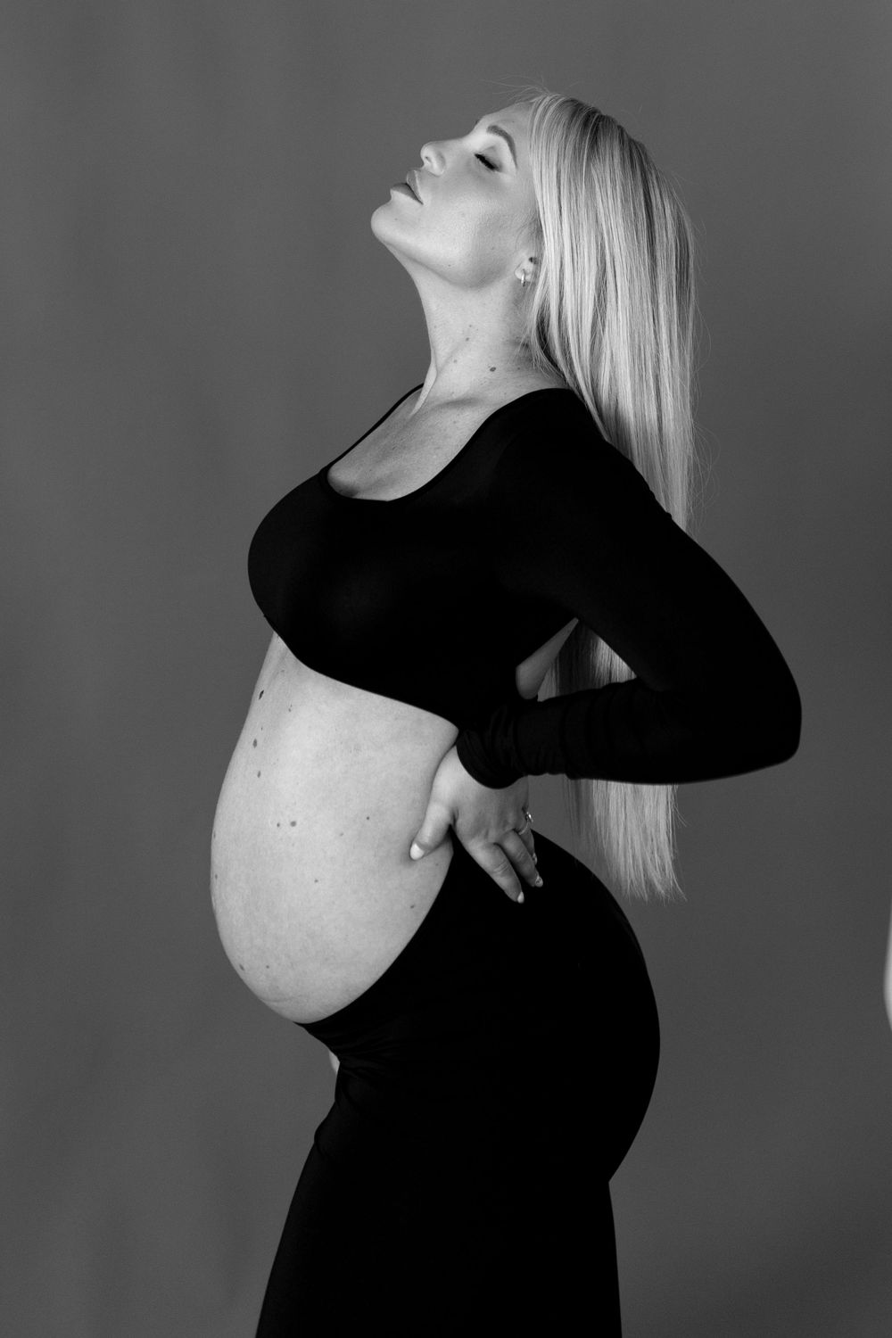 Black and white maternity portrait of a pregnant woman in a fitted black dress arching her back against gray background.