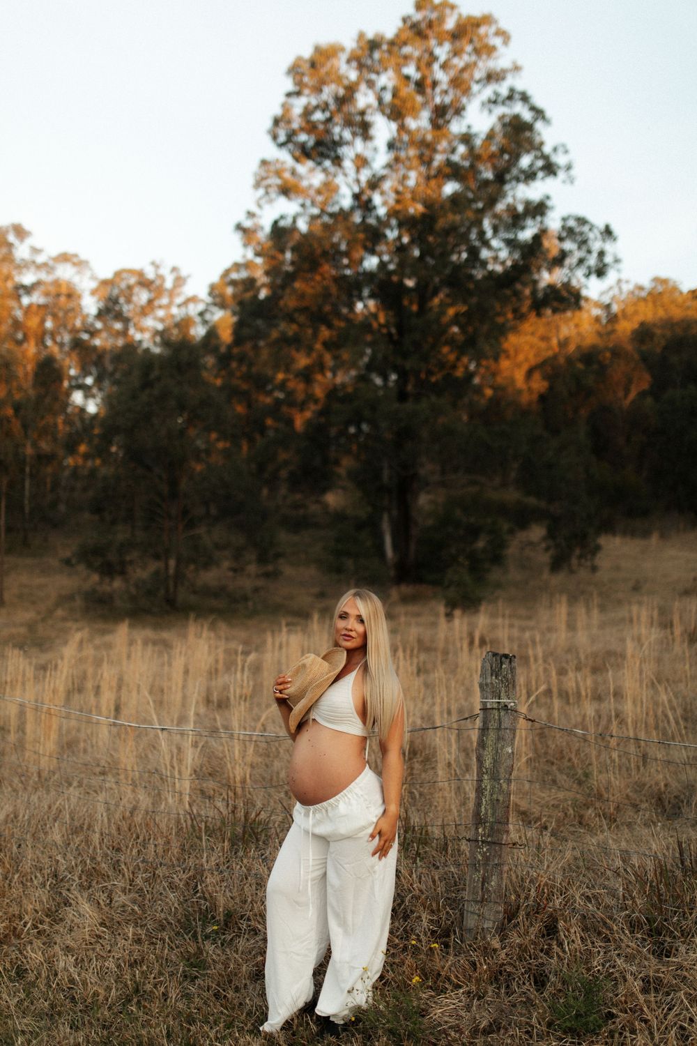 Expecting mother in white outfit poses against rustic fence and fall foliage in golden hour lighting.