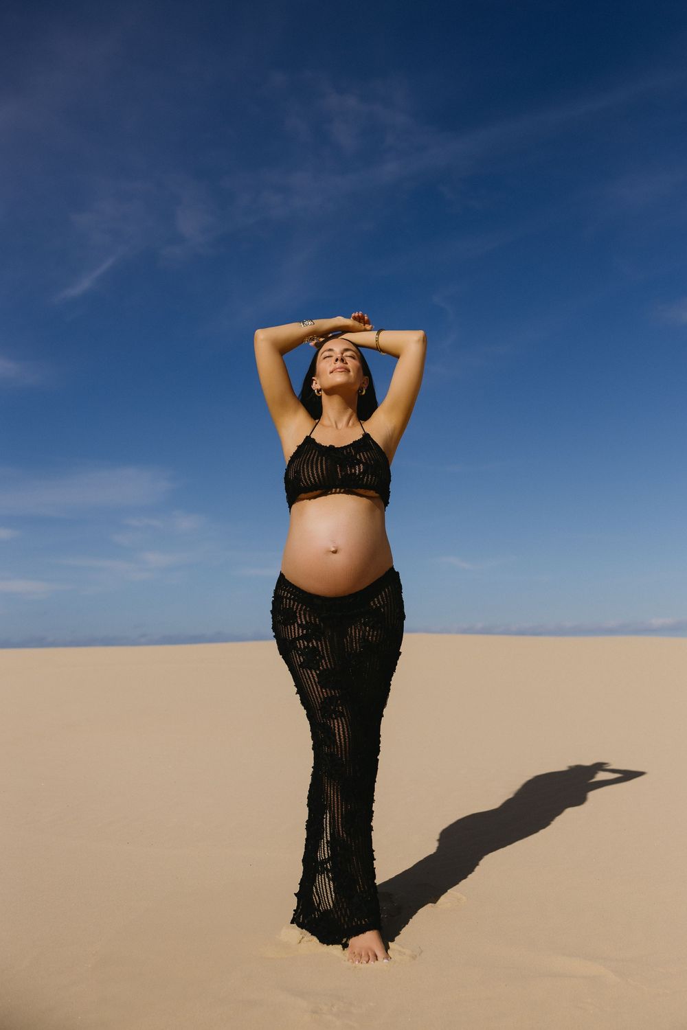 A pregnant woman in black crochet beachwear poses on sand dunes against a deep blue sky, casting a long shadow.