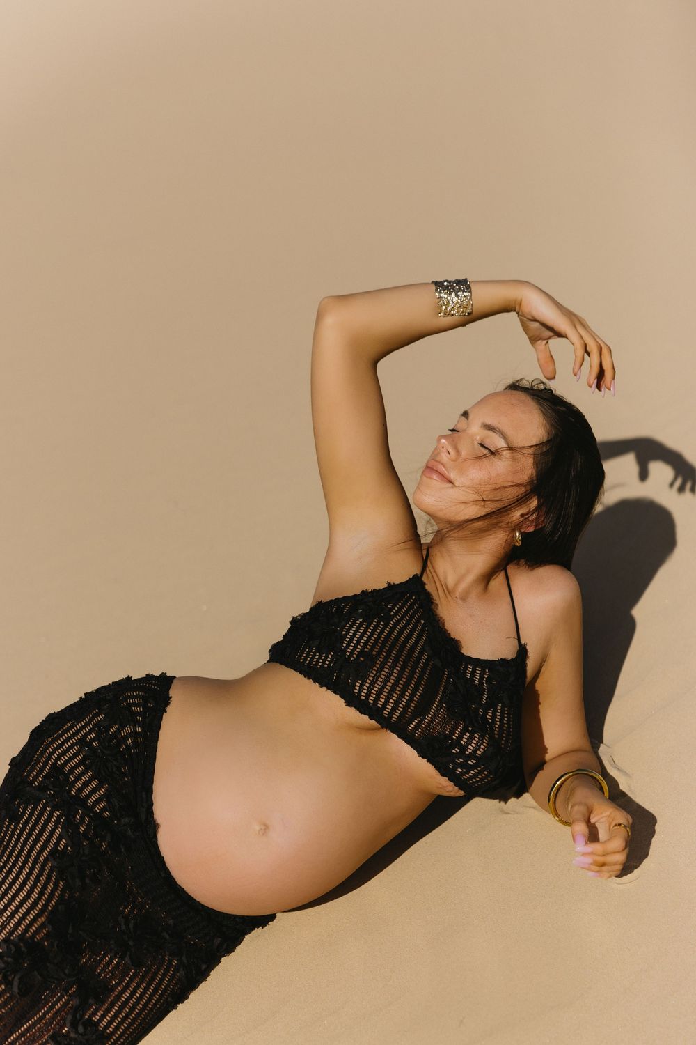 Artistic fashion photography features subject in black mesh top posing dramatically against beige backdrop with strong shadows.