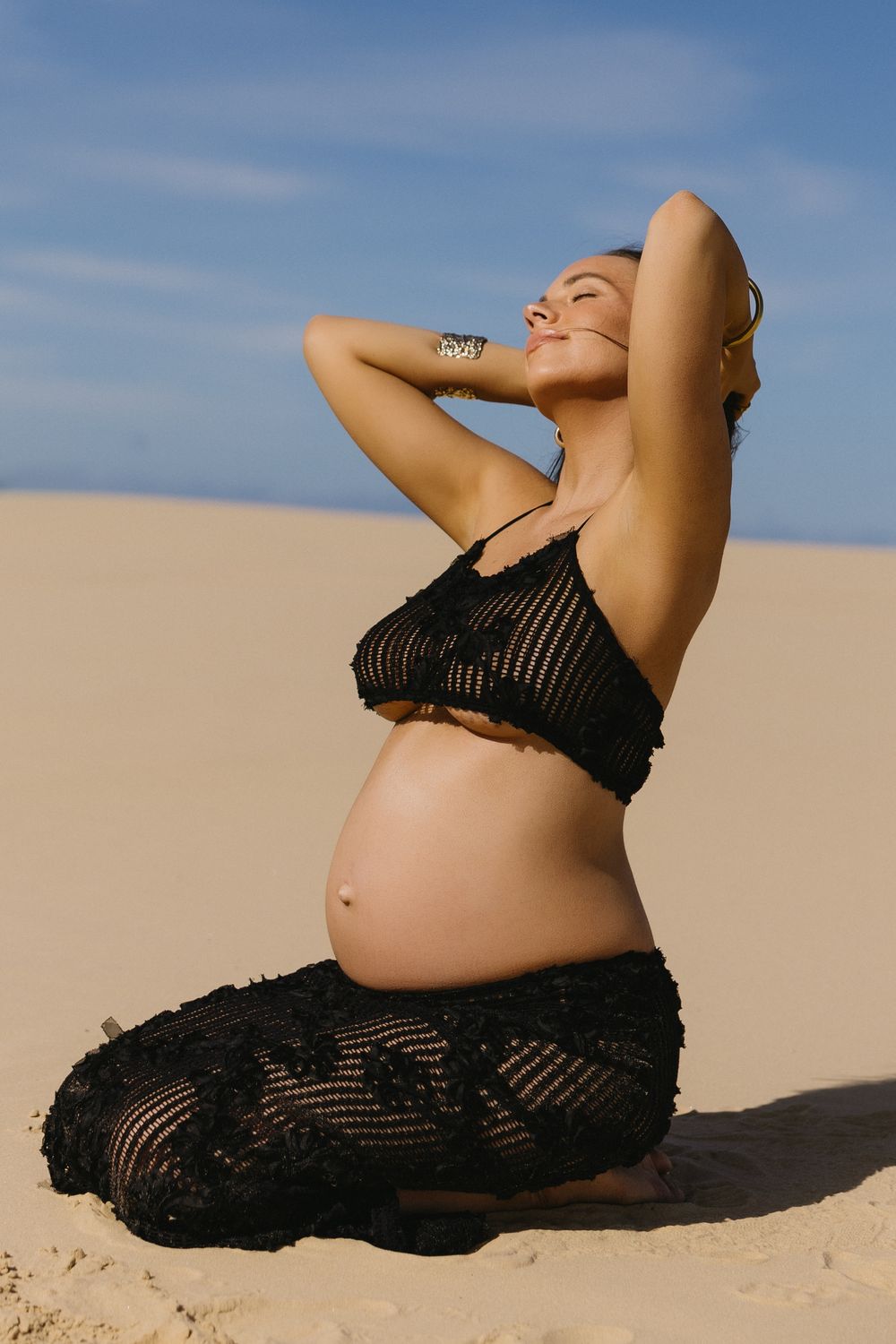 Artistic maternity photos show a pregnant woman in black lace reclining gracefully on sand dunes against a blue sky.