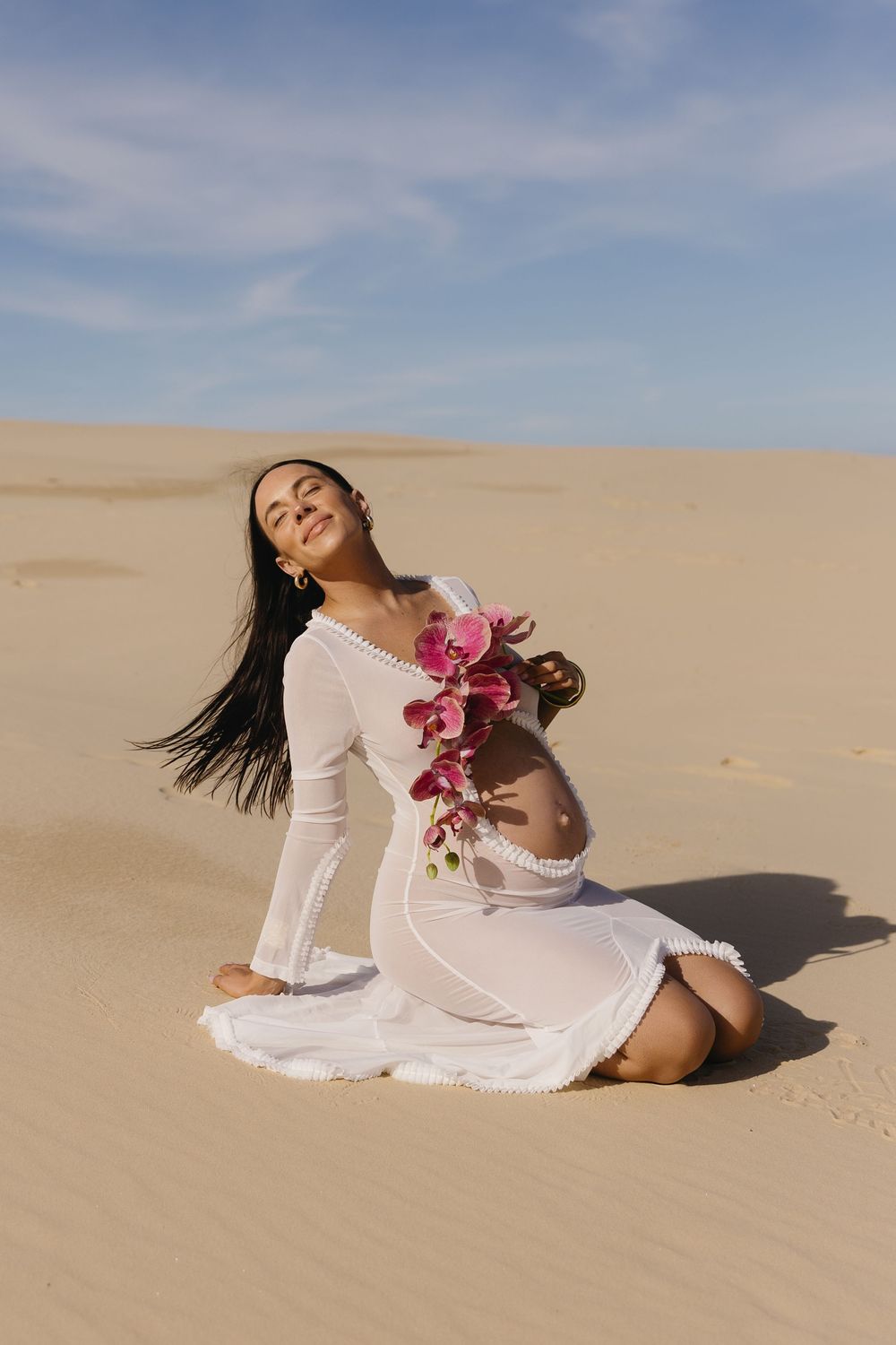 A person in a flowing white dress poses gracefully on sand dunes with pink orchids against a cloudy blue sky background.