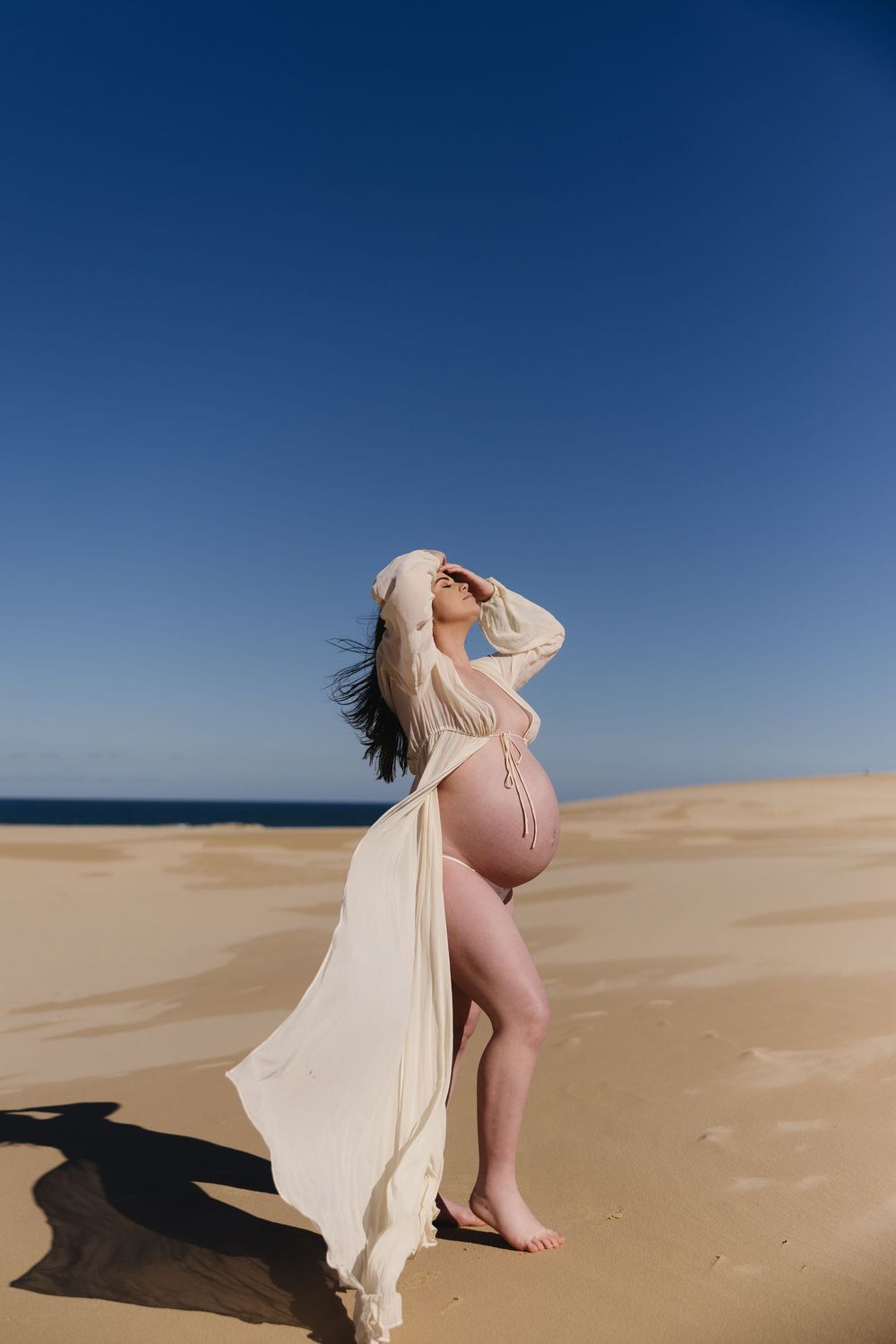 A figure in flowing sheer fabric poses dramatically against sand dunes and vivid blue sky in a desert landscape.