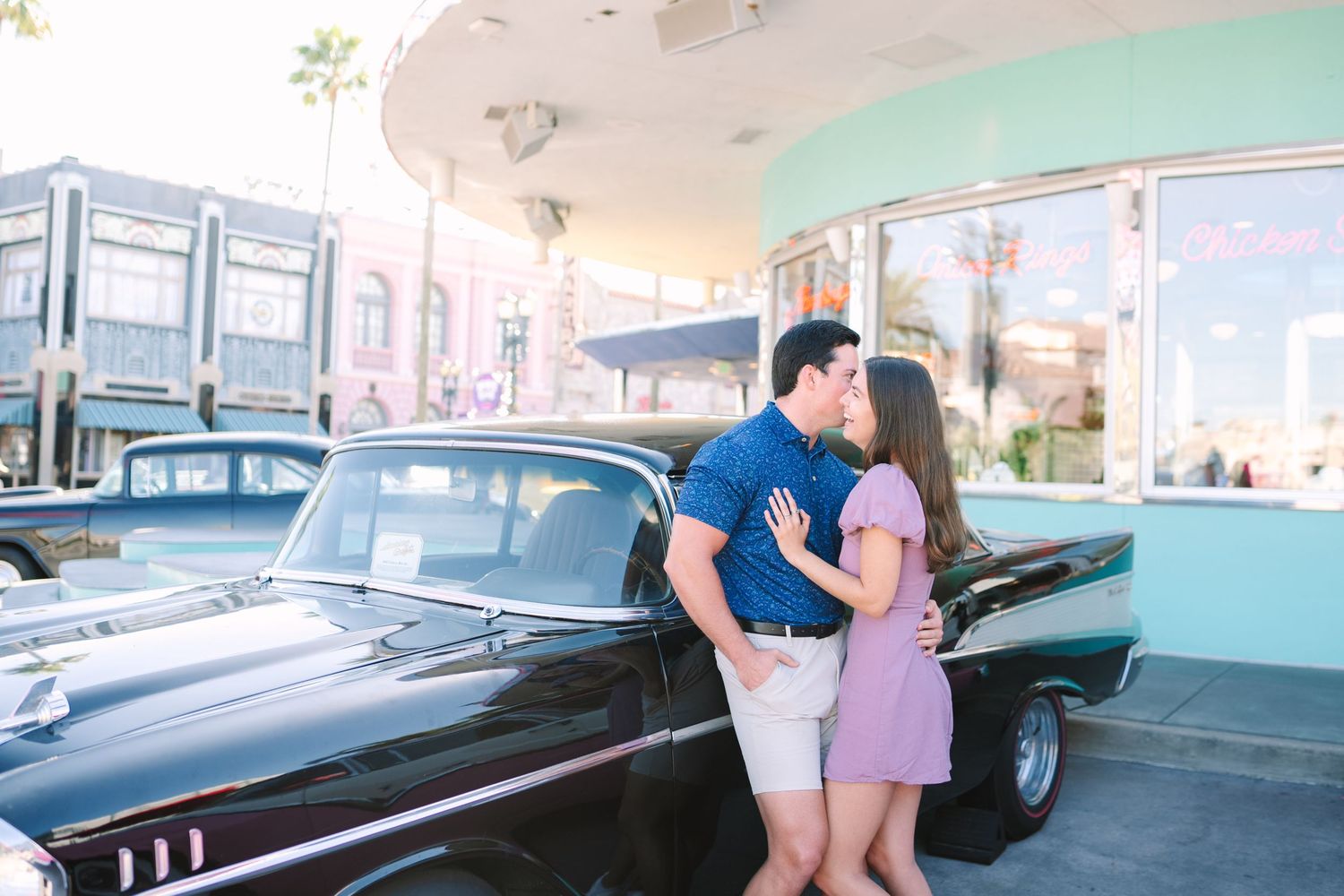 Couple kissing beside a classic black car outside a retro pink diner on a sunny day.