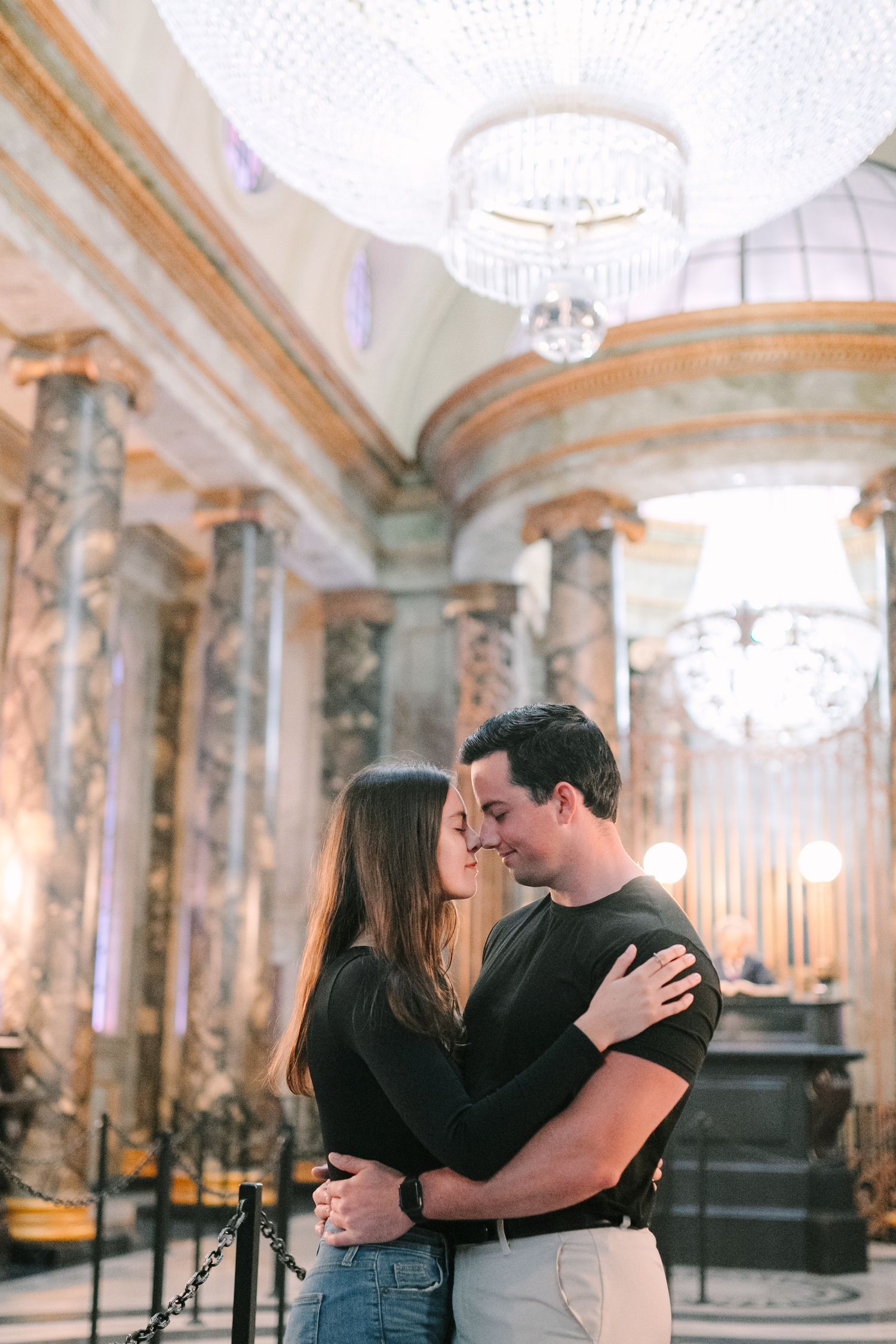 Couple sharing a romantic embrace in an elegant marble hall with crystal chandeliers and ornate columns.