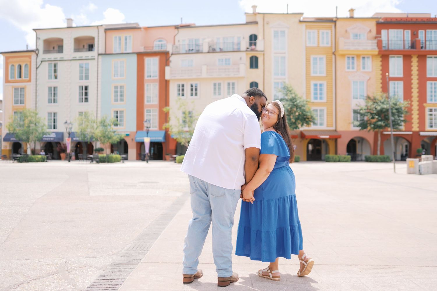 Couple sharing a romantic kiss in a colorful European-style plaza, woman in blue dress holding hands with man in white shirt.
