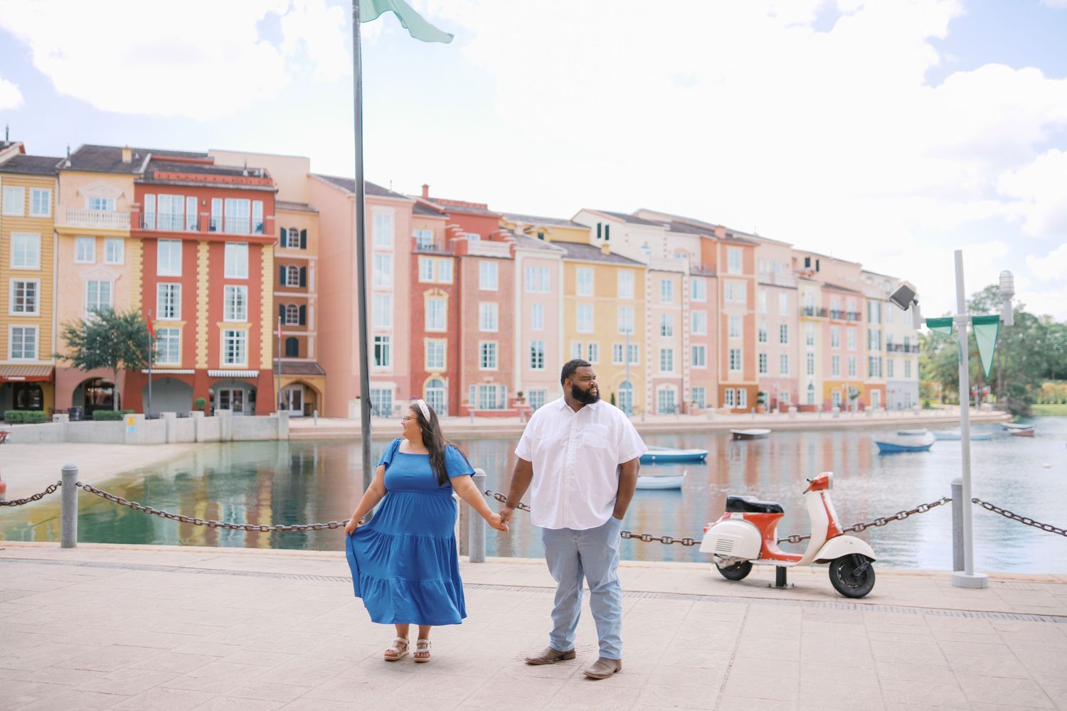 Couple holding hands on a waterfront promenade at a colorful resort with a vintage scooter nearby.