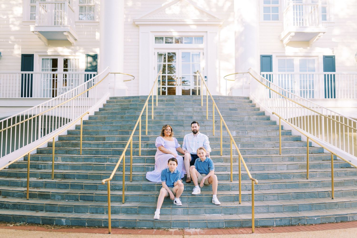 Four people sit on the steps of a grand white building with blue railings on a sunny day.