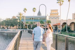 A couple walks along a scenic waterfront boardwalk with palm trees and Spanish-style architecture in the background.
