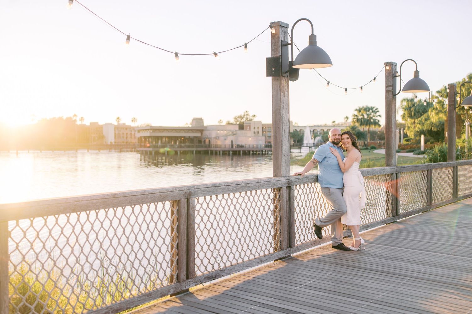 A couple embraces on a wooden boardwalk at sunset with decorative lamp posts and waterfront views in the background.
