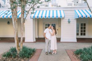 Couple in light colored outfits poses together in front of a white building with blue and white striped awnings.