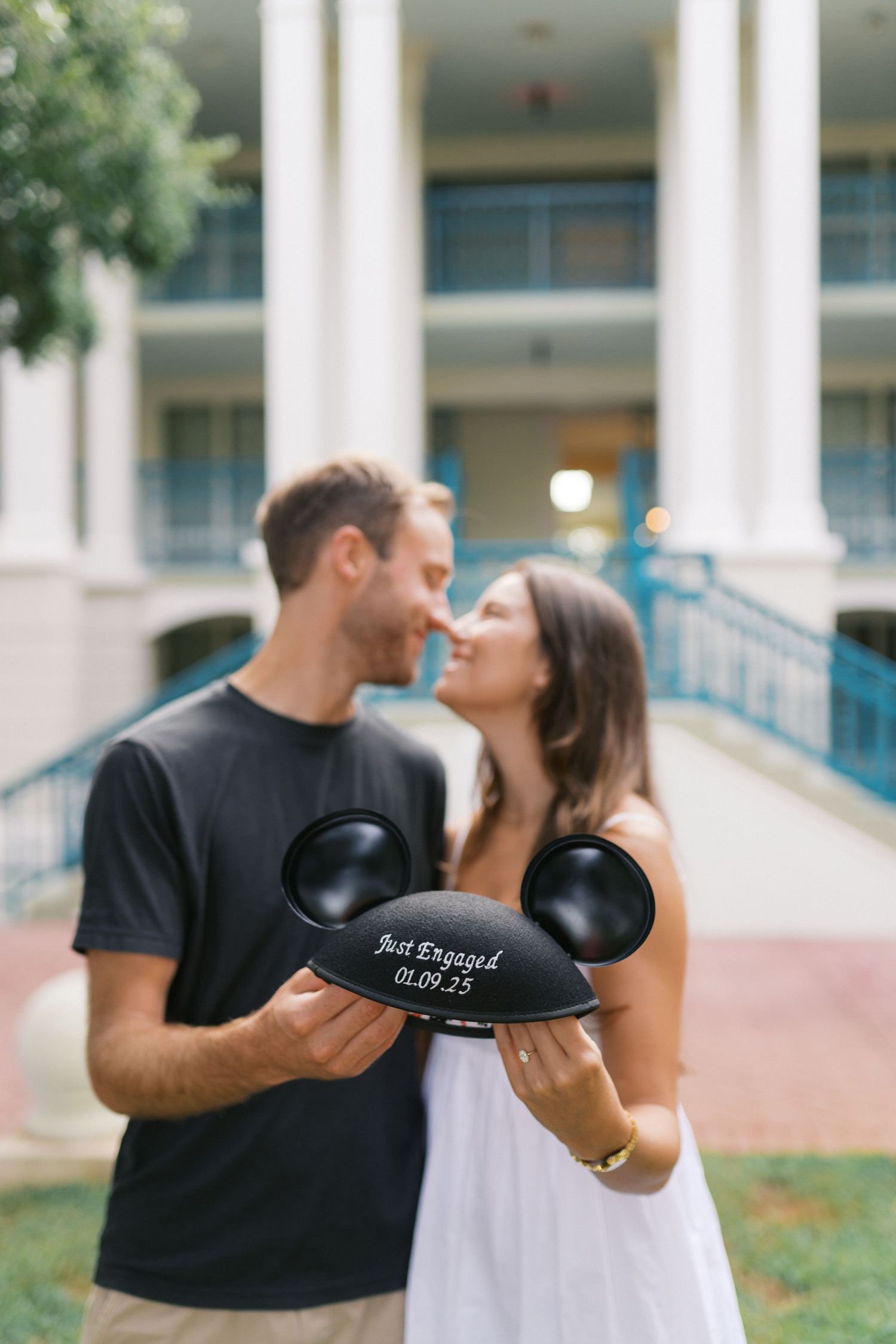 A couple shares a romantic moment while holding a Mickey Mouse ear hat at a Disney resort with white columns in background.
