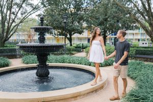 A couple stands near an ornate garden fountain surrounded by lush landscaping and trees in a peaceful courtyard setting.