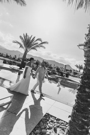 Wedding couple walking by pool with palm trees and mountains in background.