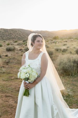 A bride in a classic white wedding dress holds a bouquet of white flowers in a desert landscape at sunset.