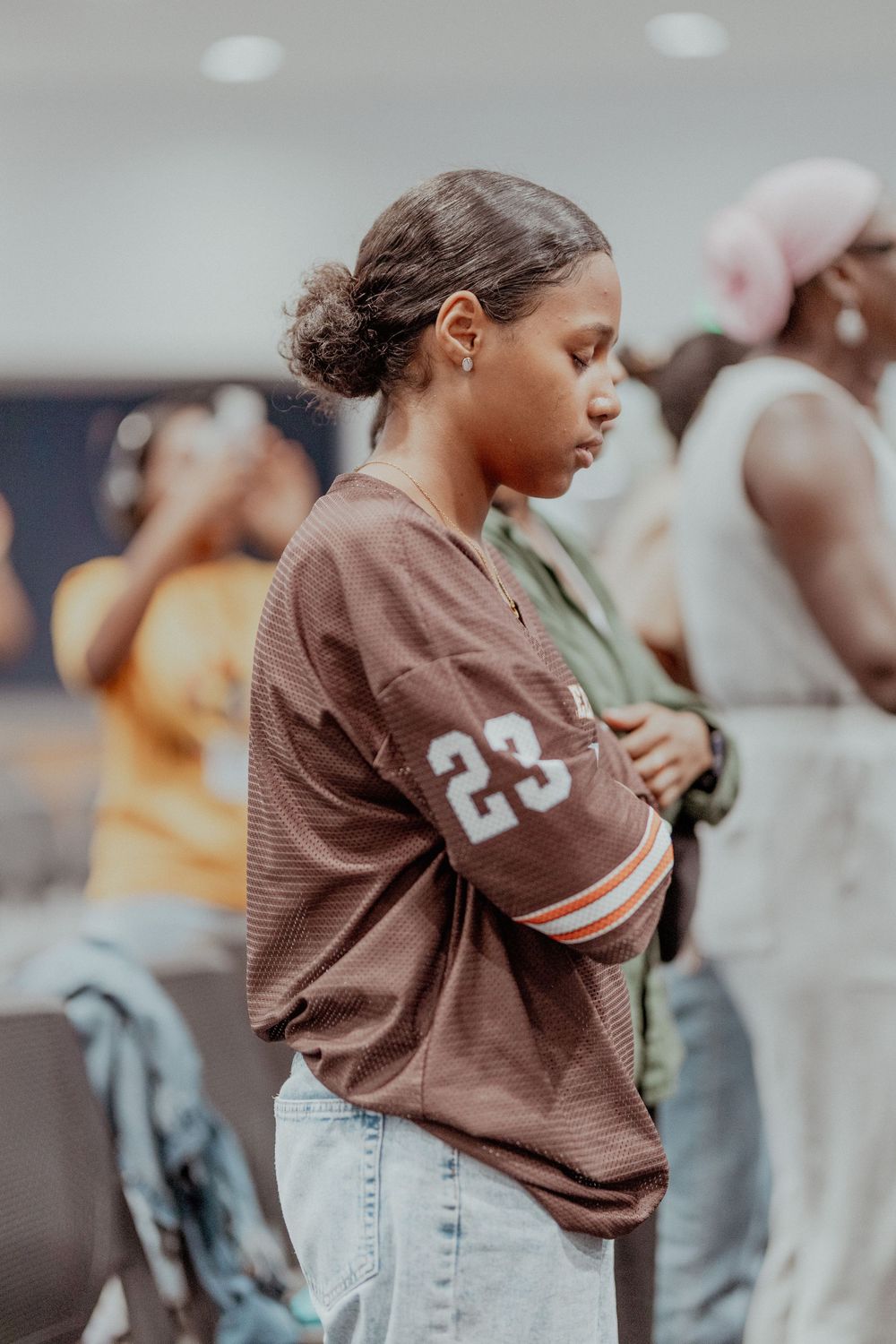 A person wearing a brown jersey and light denim jeans stands in a casual pose against a blurred background.