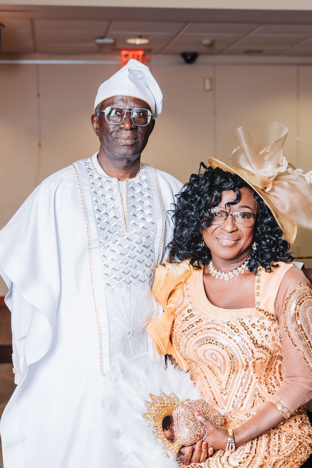 Two people in formal attire wearing traditional West African clothing in white and peach colors with elegant accessories.