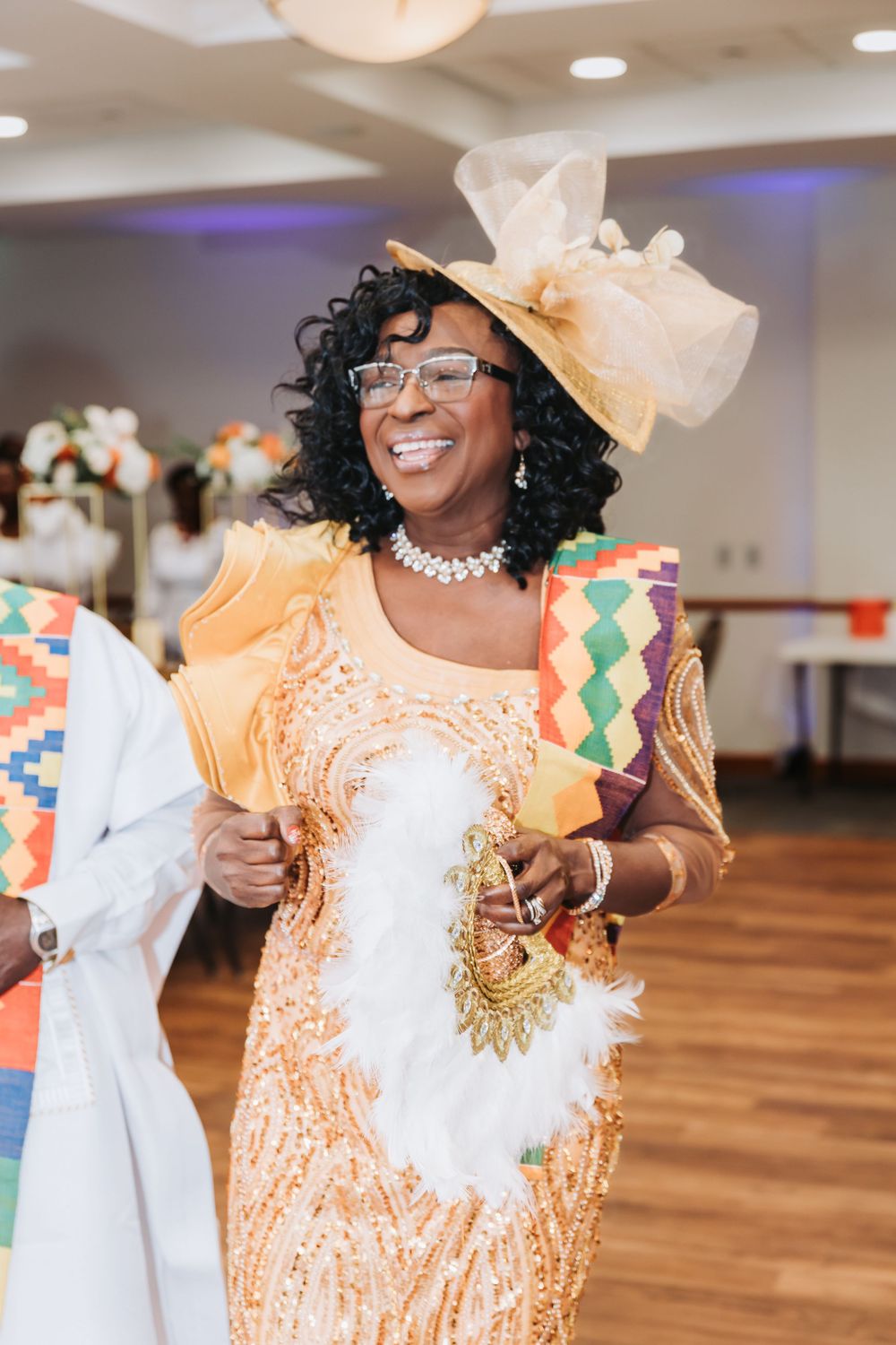 A person wearing a peach-colored dress and white fascinator hat smiles joyfully at an indoor celebration.