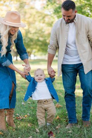 Couple in casual denim outfits helping a happy toddler walk on grass in a sunny outdoor setting.