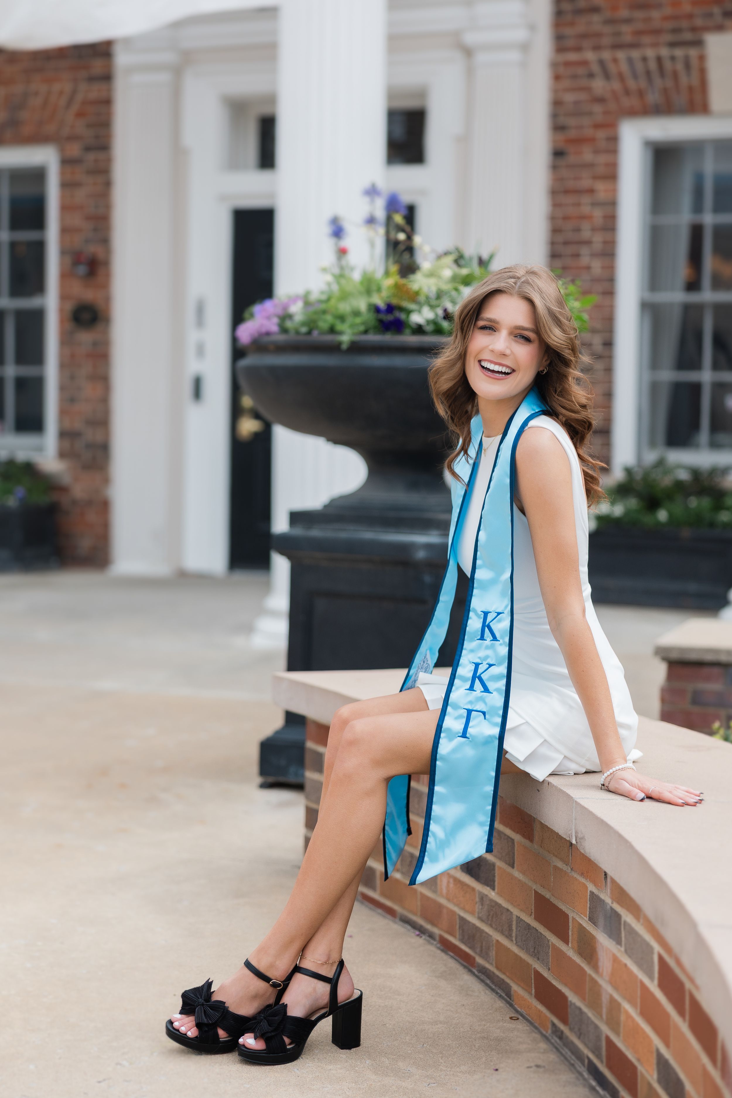 A graduate in a light blue sash and white dress sits casually on a brick ledge near a columned building entrance.