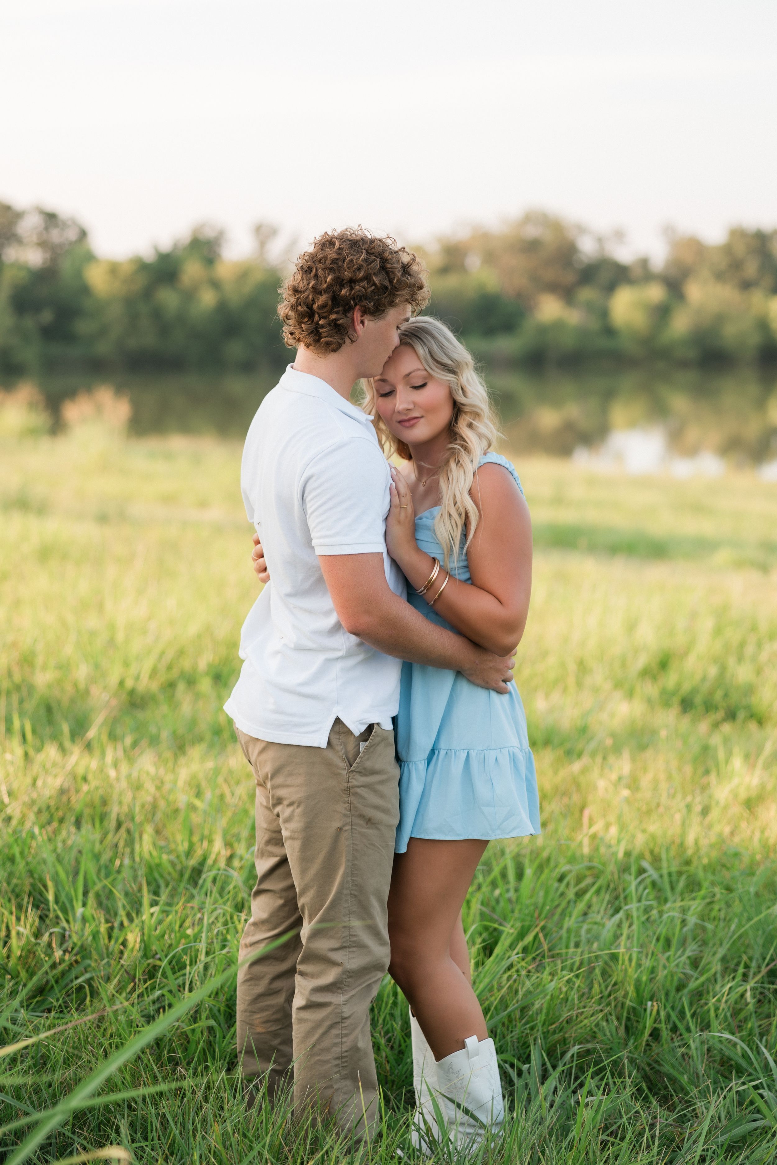 A romantic couple embraces in a sunlit meadow with tall grass and trees in the background during golden hour.