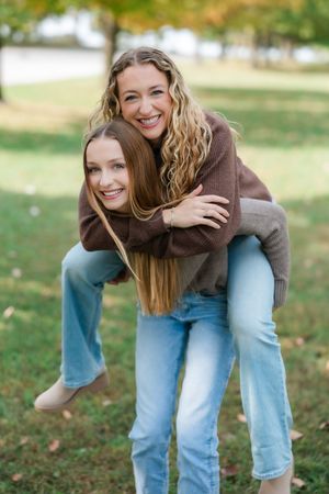 Two friends share a playful piggyback moment outdoors while wearing brown sweaters and blue jeans on an autumn day.