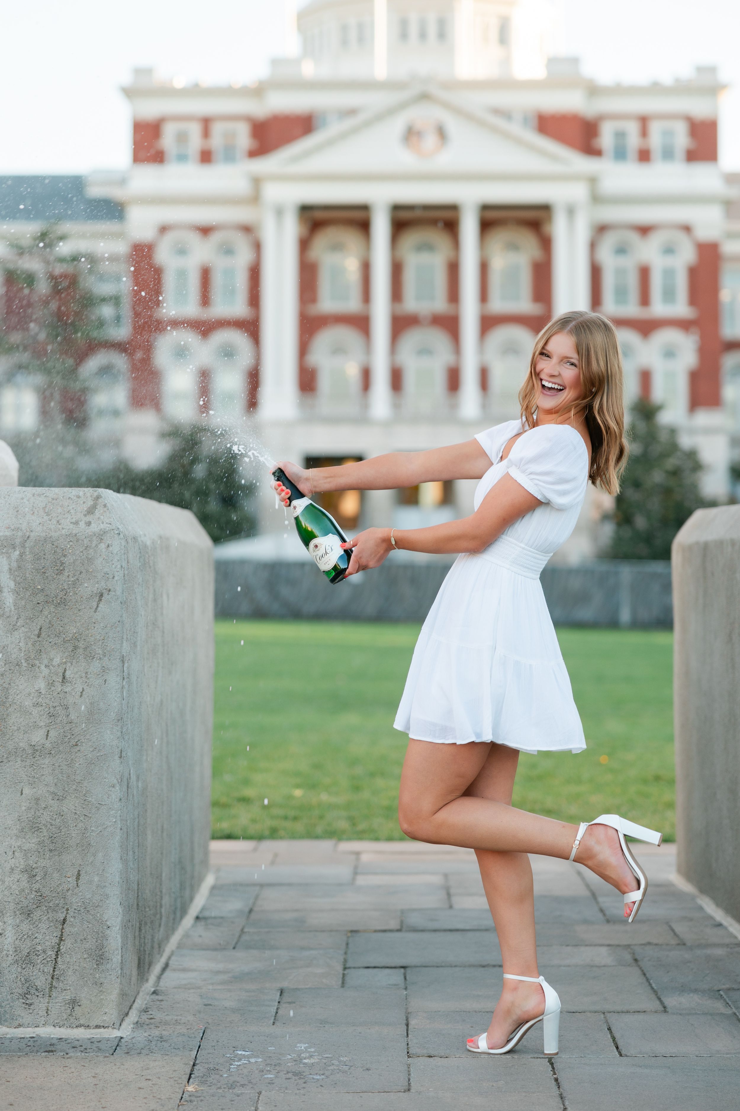 A person in a white dress and heels poses playfully with a champagne bottle in front of a grand historic building.